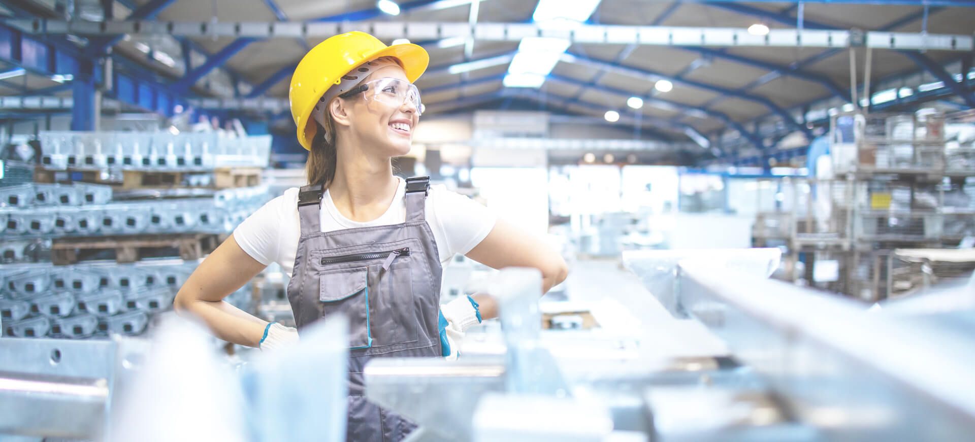 Lächelnde Mitarbeiterin mit gelbem Schutzhelm in einer Produktionshalle